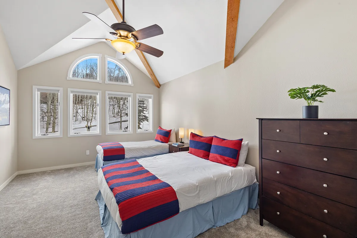 A welcoming bedroom with two beds featuring striped bedding, a wooden dresser, ceiling fan, and large windows that fill the room with natural light. The setup is ideal for families or groups, offering comfort and flexibility.