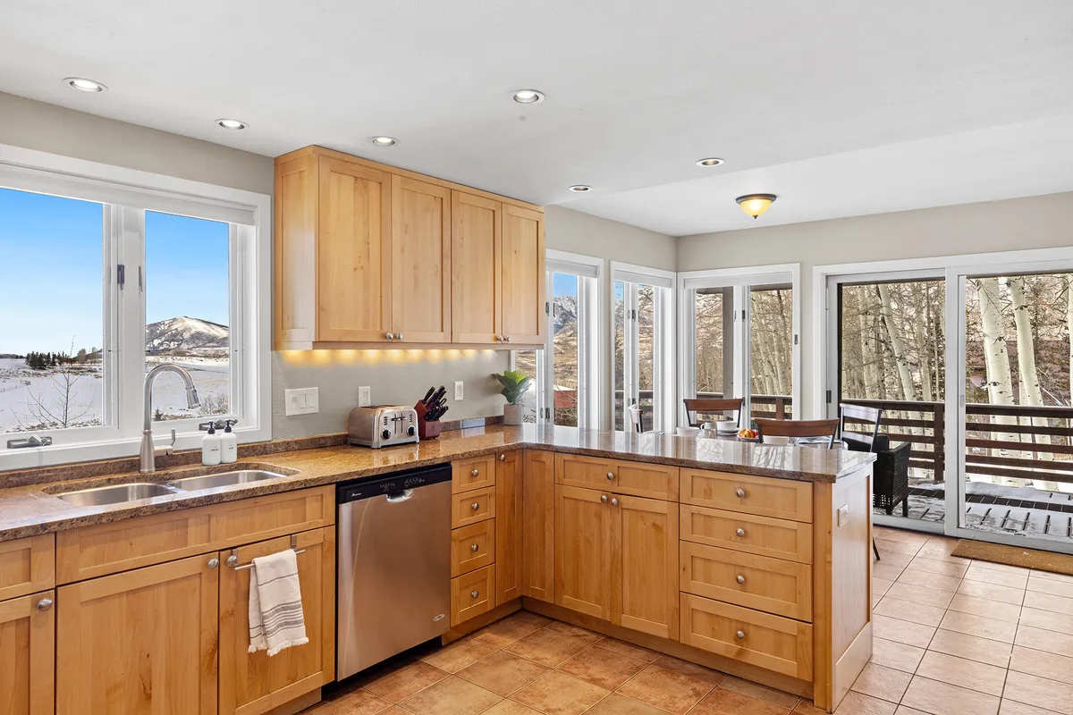 This image features a spacious kitchen with a central island, gleaming stainless steel appliances, large windows filling the space with natural light, and a granite countertop. The scene suggests a space designed for both culinary creativity and social gatherings, with ample prep room and inviting ambience.