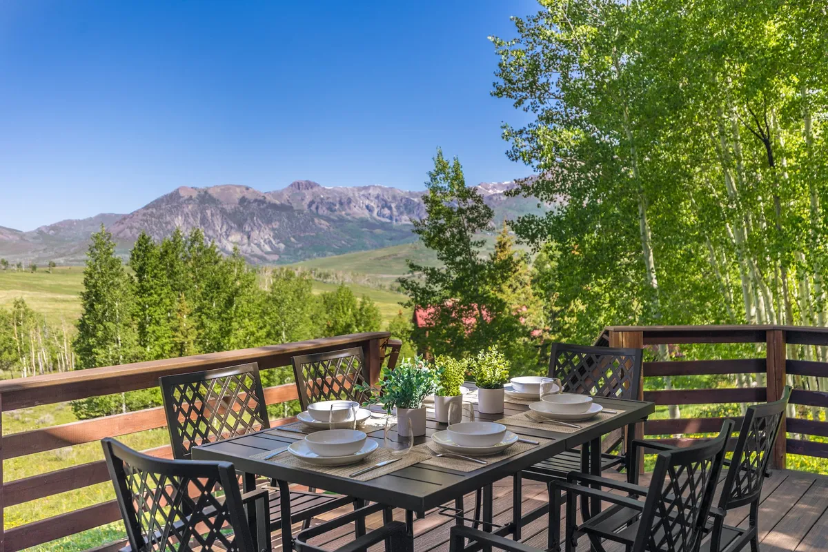 An exterior dining area set for a meal, surrounded by lush green foliage and overlooking panoramic mountain views. The space is inviting and ideal for gatherings, with natural light and abundant seating.