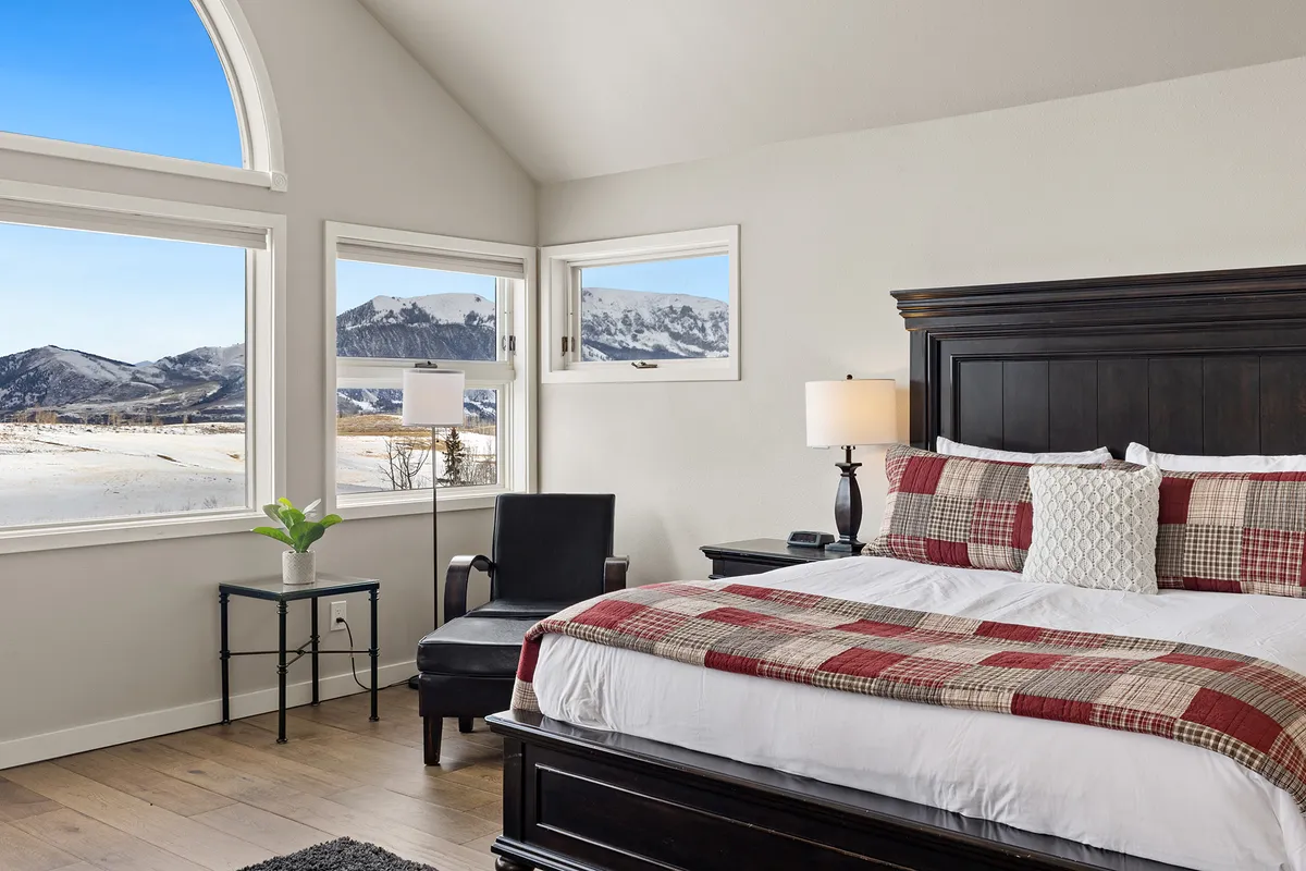 A cozy master bedroom featuring a king-sized bed with plaid bedding, an armchair in the corner, and expansive windows providing breathtaking mountain views. The room is filled with natural light, giving it a serene and peaceful atmosphere.
