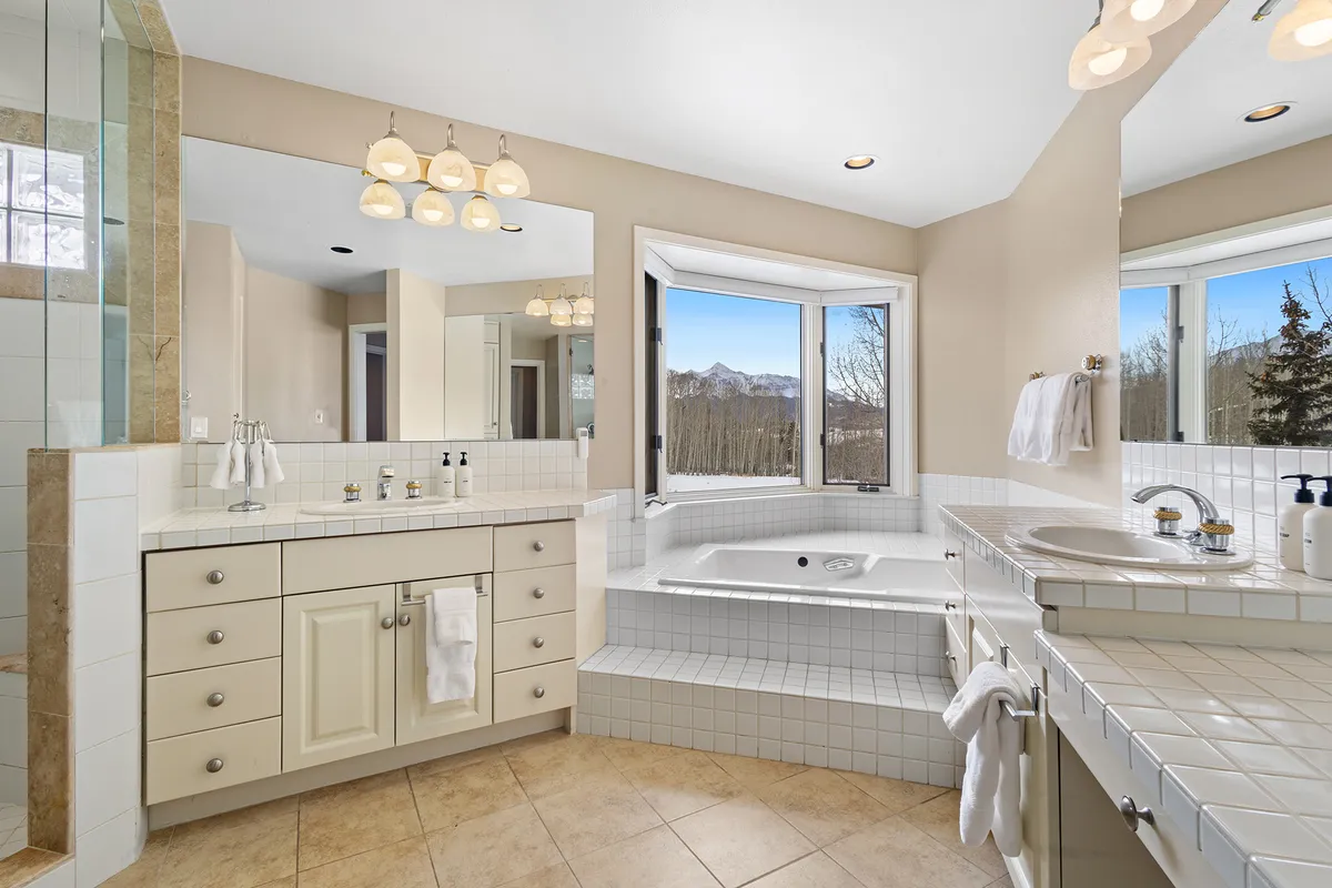 A luxurious bathroom showcasing a jacuzzi tub positioned beside large windows that frame stunning mountain views, complemented by a double sink vanity. The space feels calm, refreshing, and spa-like.