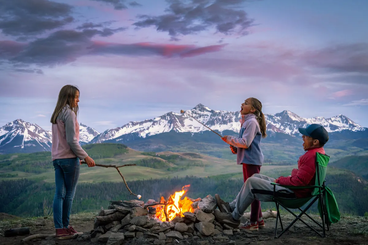 A joyful family scene by a campfire, with children roasting marshmallows and an adult supervising. The mountains provide a scenic backdrop, and the group appears engaged in storytelling and laughter.