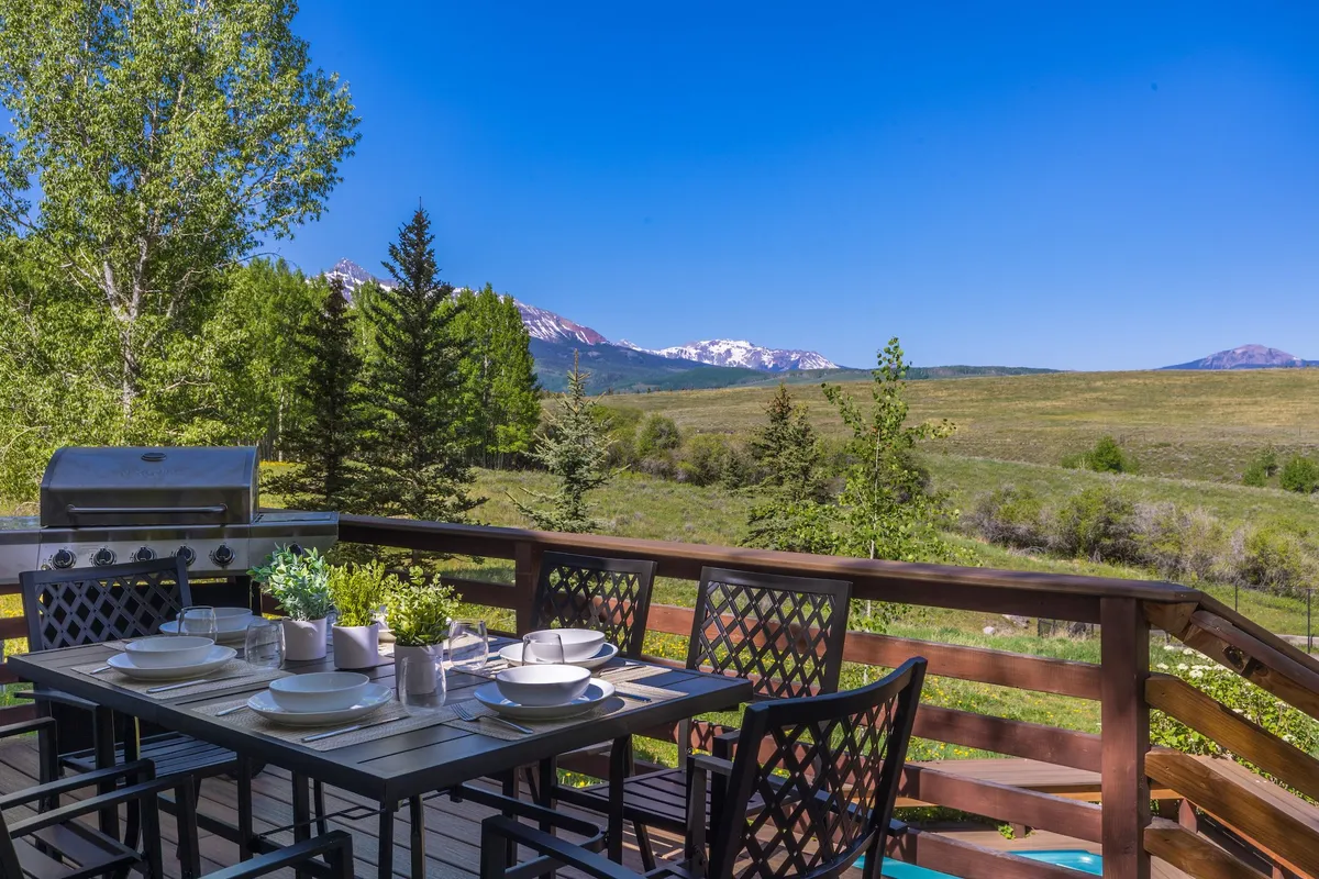 An outdoor deck set for a meal, featuring a grill, dining table, and mountain views bordered by lush green trees. The scene evokes a relaxed, cheerful vibe perfect for barbecues, morning coffee, or starlit gatherings.