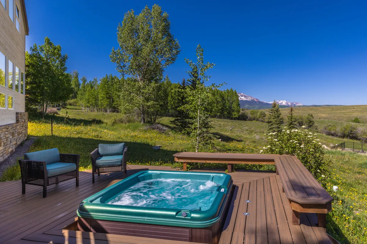 A hot tub sits on a wooden deck surrounded by trees, with expansive mountain views in the background. The tranquil setting shows the perfect spot to relax after a day of hiking or attending local festivals, with the fresh mountain air adding to the rejuvenating atmosphere.