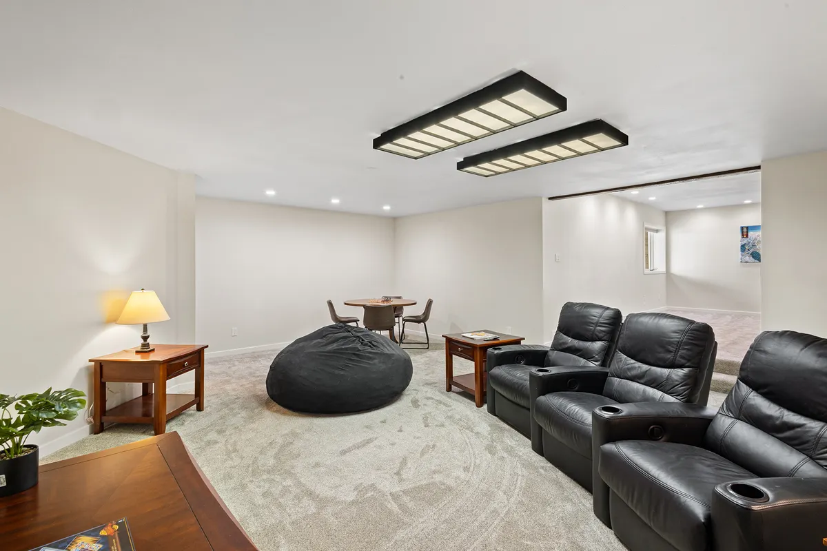 A casual basement entertainment area featuring reclining chairs, a bean bag chair, a small table with chairs, and a table lamp. The space is arranged for relaxation and group activities, with a focus on comfort and versatility. It exudes a cozy atmosphere perfect for movie nights or casual gatherings.