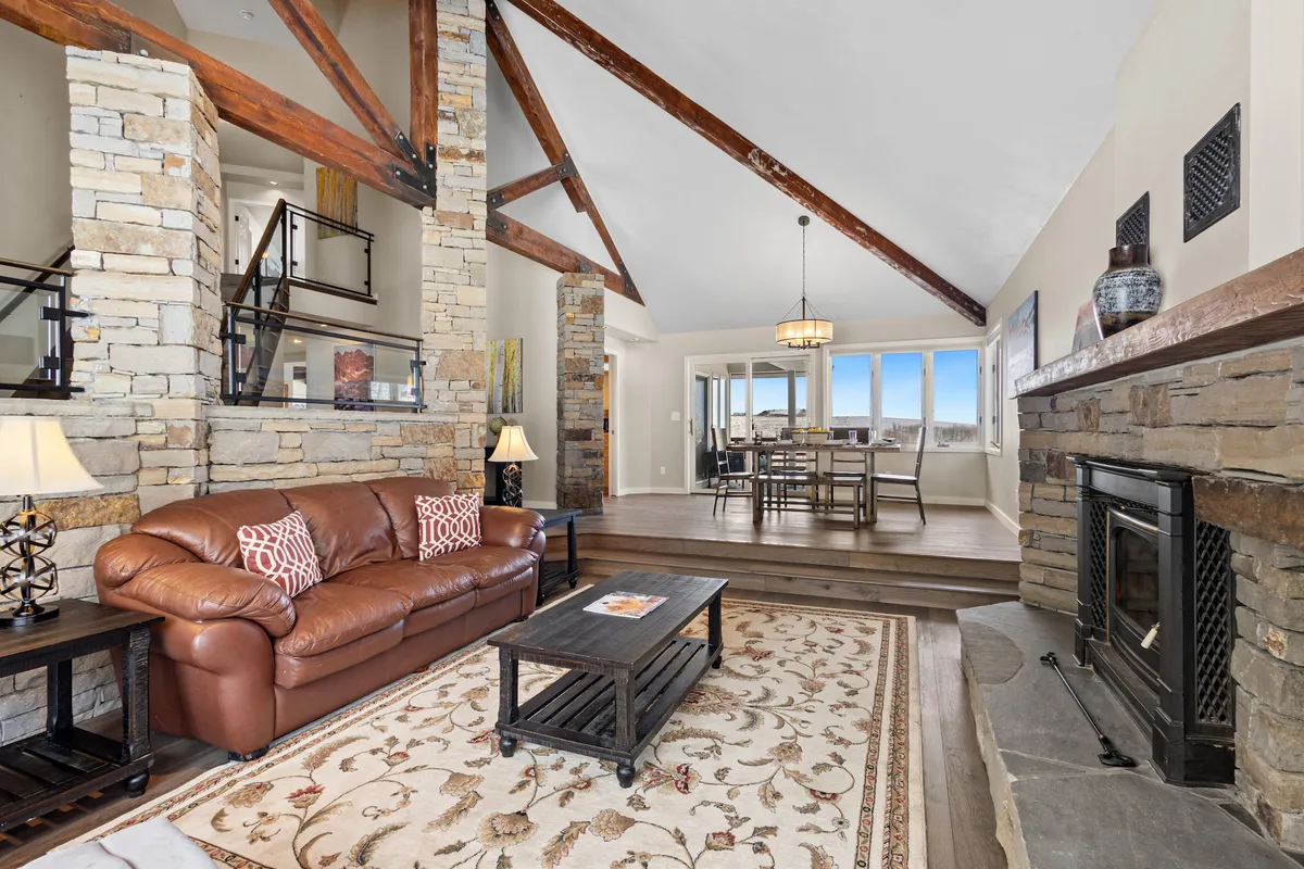 A warm, open-concept living and dining area featuring a leather sofa in front of a rustic stone fireplace, exposed wooden beams, and a dining area with large windows that frame mountain views. The sunlight pours in, accentuating the cozy, inviting mood perfect for gathering and relaxing after outdoor adventures.