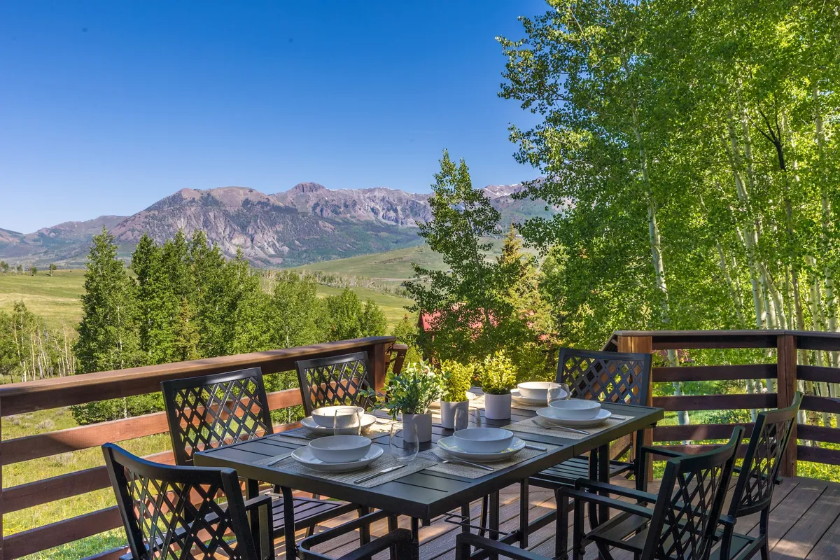 An outdoor dining area with a table set against a panoramic mountain view and surrounded by lush greenery. The space invites guests to enjoy meals or drinks while immersed in the natural landscape.